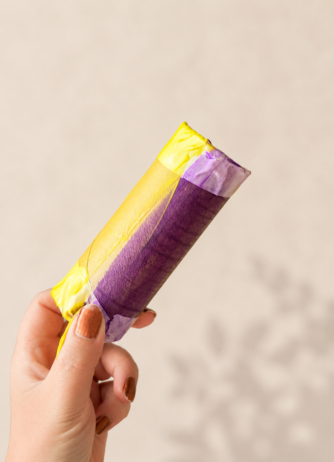 Hand holding a purple and yellow painted toilet paper roll shaker sealed with clear tape